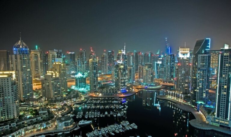 Panorama miasta Dubaj nocą z widokiem na zatokę i oświetlone wieżowce oraz jachty w marinie, widoczne budynki mieszkalne i biurowe.