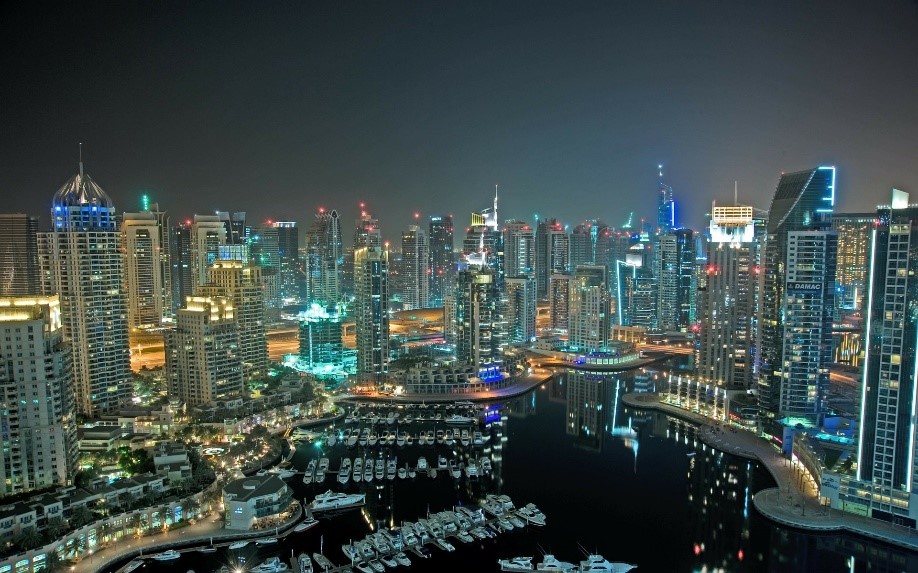 Panorama miasta Dubaj nocą z widokiem na zatokę i oświetlone wieżowce oraz jachty w marinie, widoczne budynki mieszkalne i biurowe.