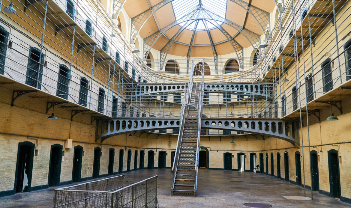 Wnętrze więzienia Kilmainham Gaol w Dublinie z metalowymi galeriami, celami i centralną klatką schodową pod przeszklonym dachem.
