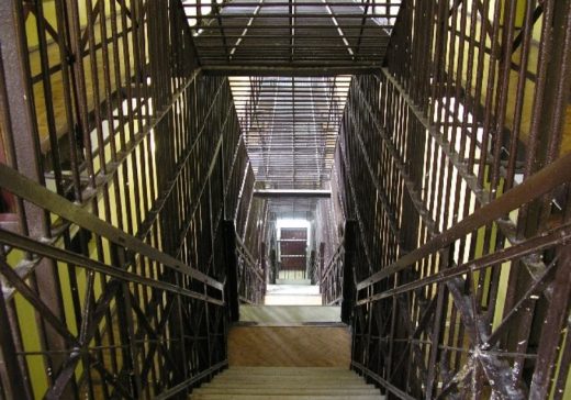 Wąska klatka schodowa z metalowymi balustradami i kratami, widok w dół na schody i kolejne kondygnacje z metalowymi zabezpieczeniami po obu stronach.